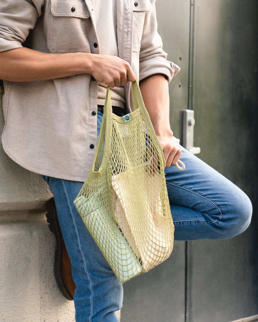 Person holding a green string bag with items, leaning on a concrete surface.