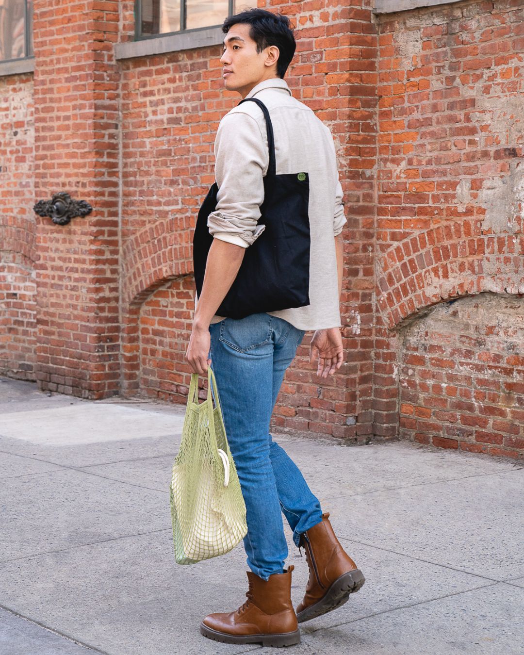 Man standing against a brick wall wearing a white shirt, blue jeans, brown boots, and carrying a green string bag.