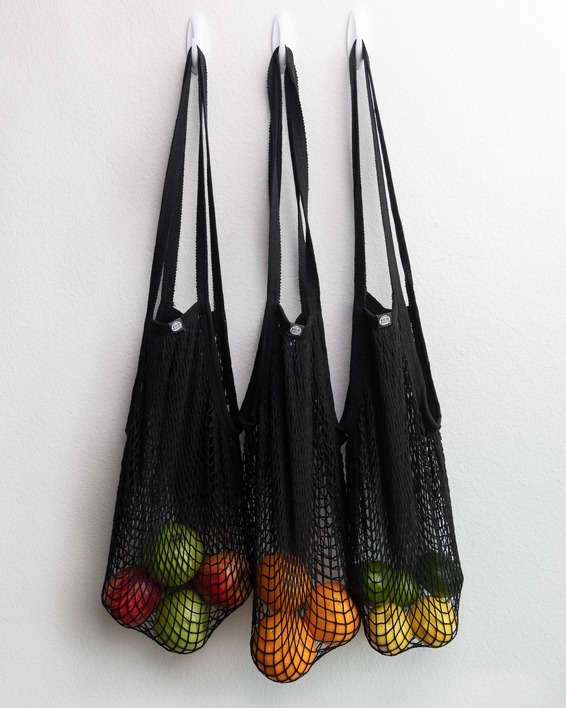 Three black mesh reusable cotton string bags filled with fruits on a white background