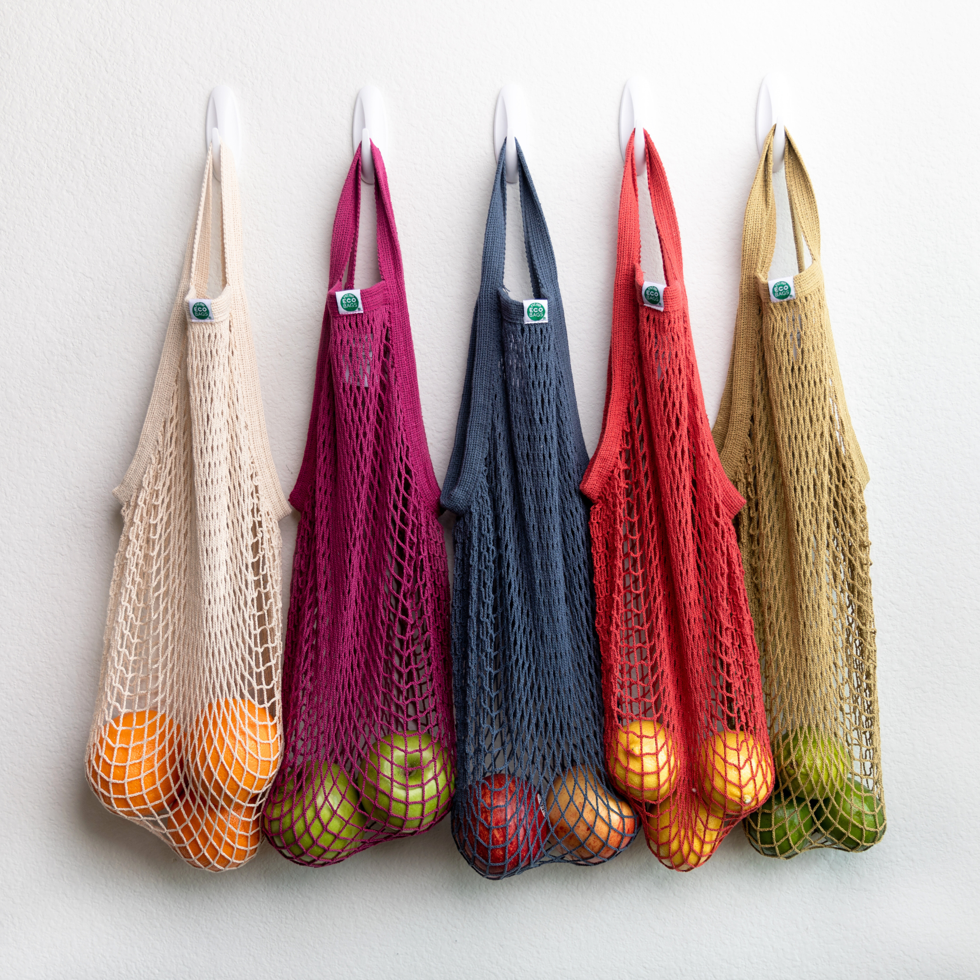 Five tote handle string bags in different earth tone colors, hanging on a wall, filled with produce.