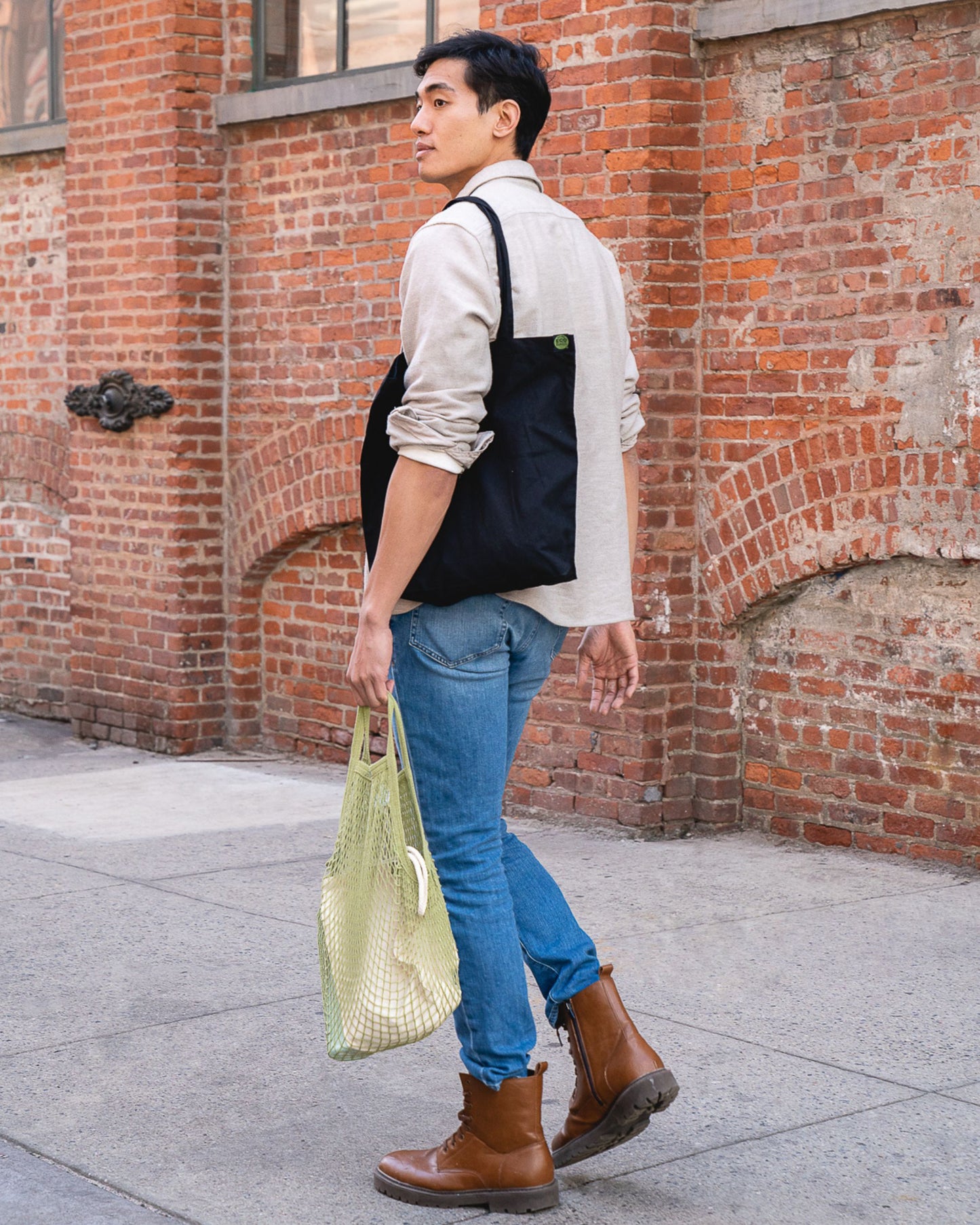 Man holding a green string bag and wearing a black tote bag against a brick wall.