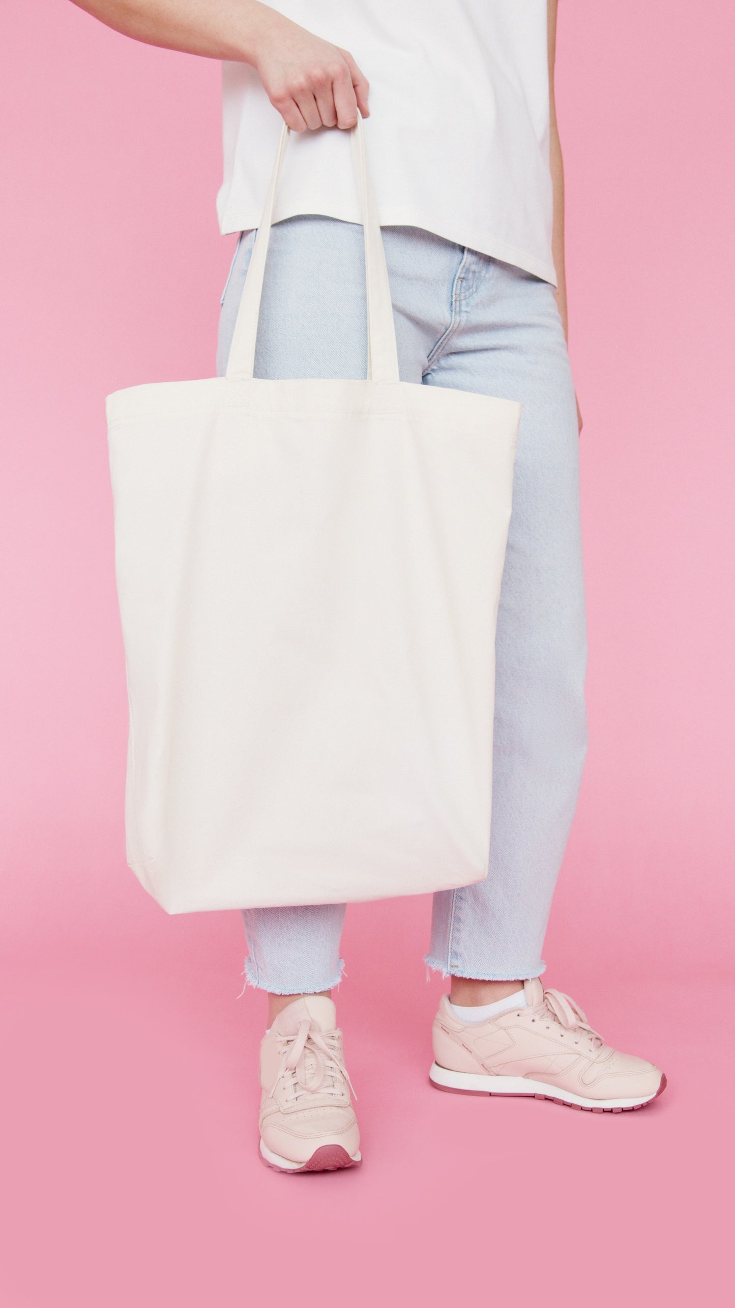 Person holding a natural organic canvas book tote bag, standing against a pink background.