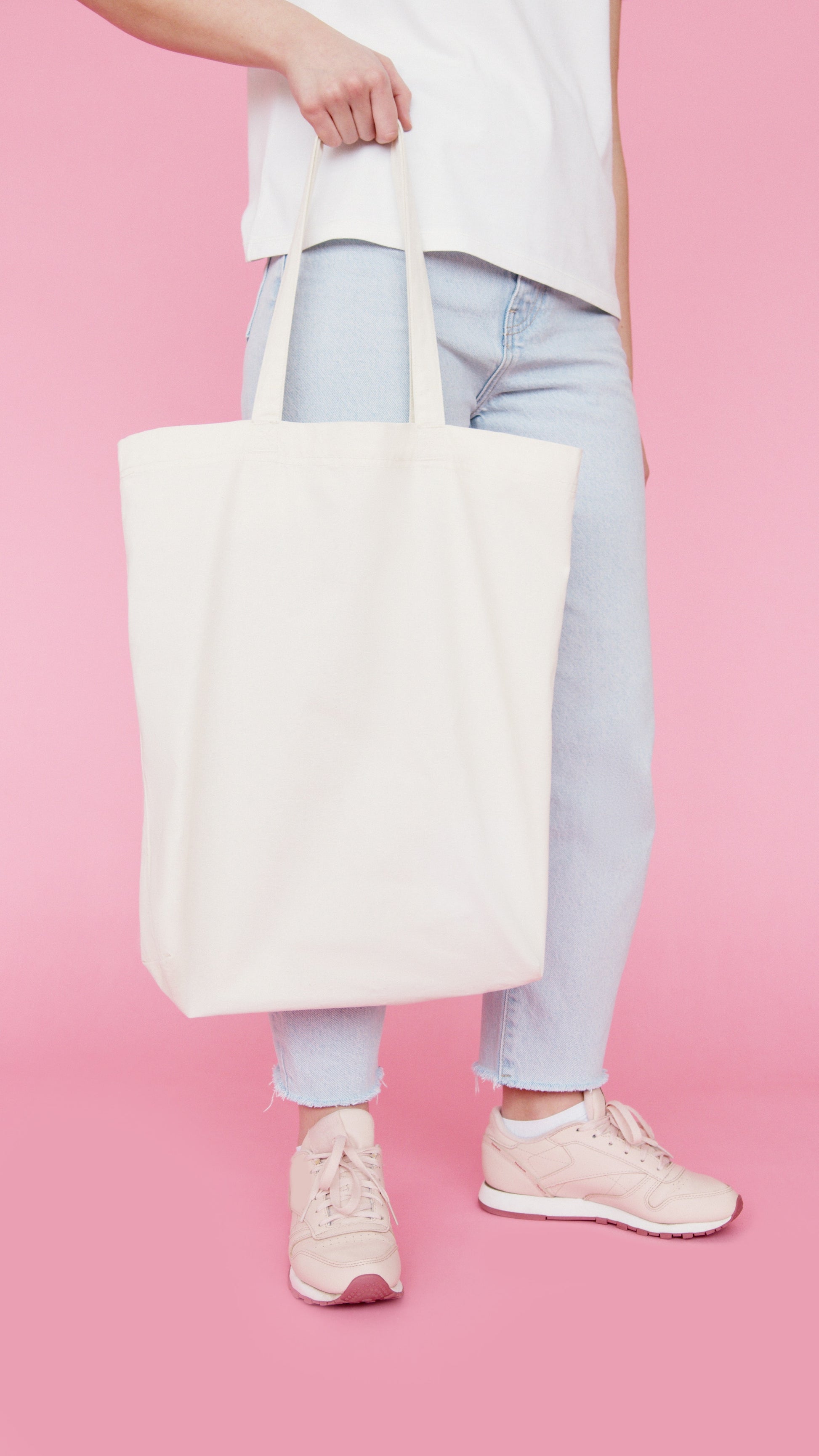 Person holding a natural organic canvas book tote bag, standing against a pink background.