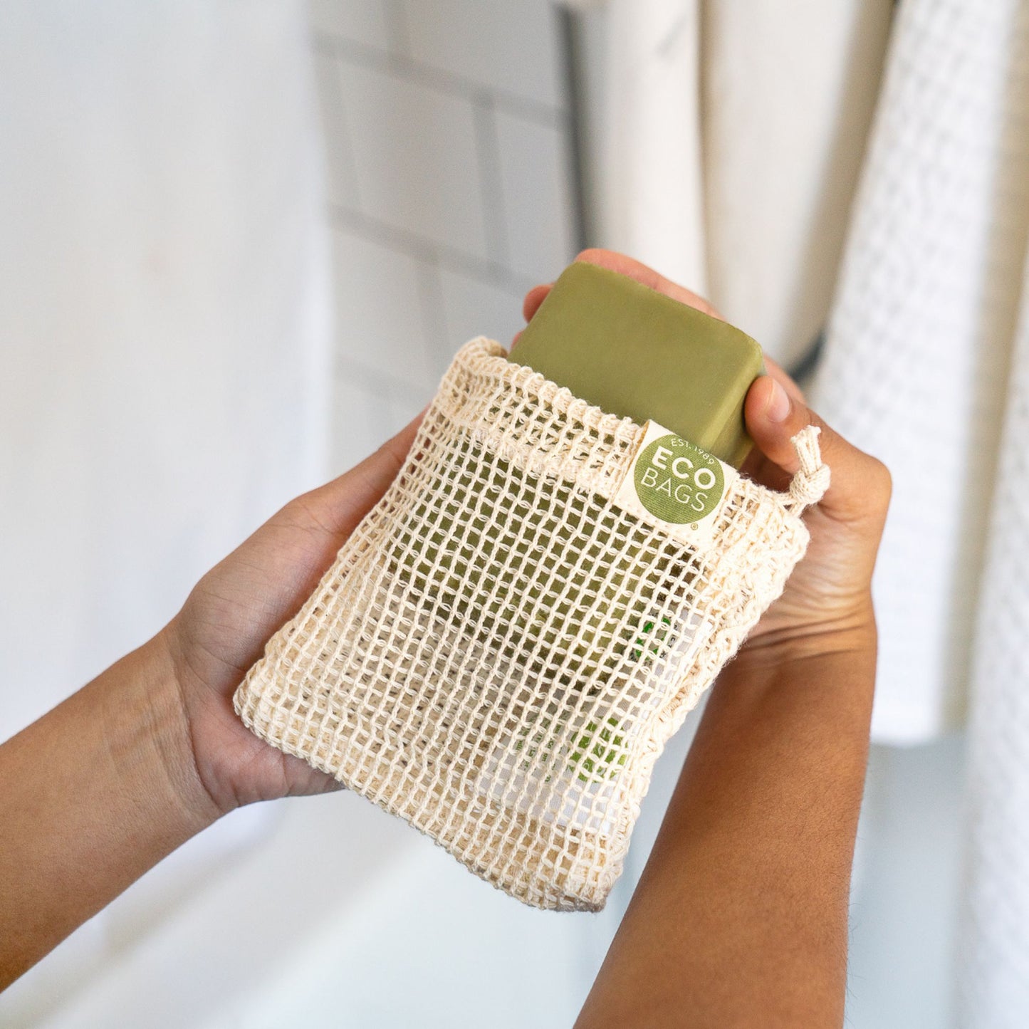 Hands hold an ECOBAGS organic cotton Soap Saver Bag containing a green bar of soap