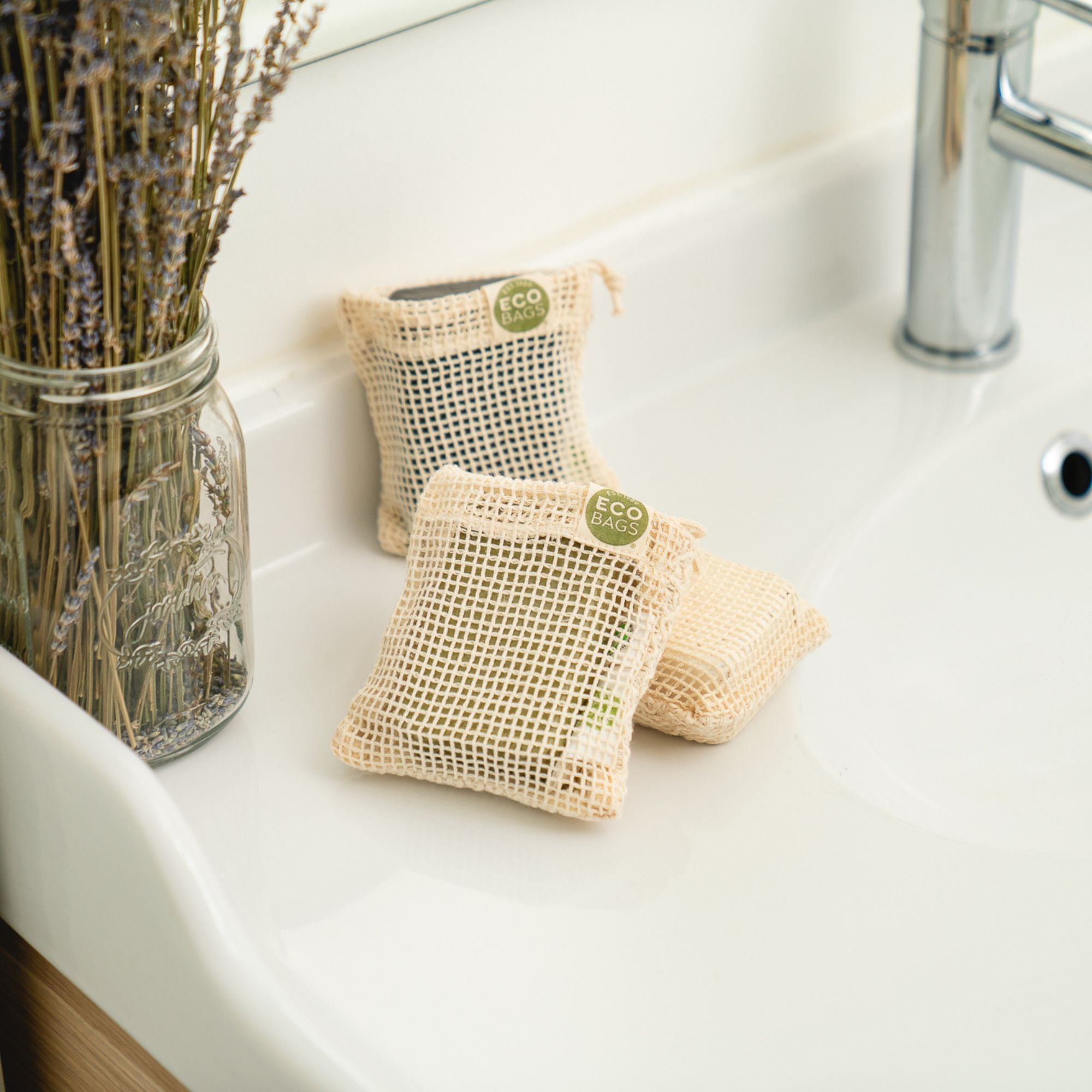 Three ECOBAGS Soap Saver Bags made from natural, untreated organic cotton sit on a white sink next to lavender in a Mason jar. 
