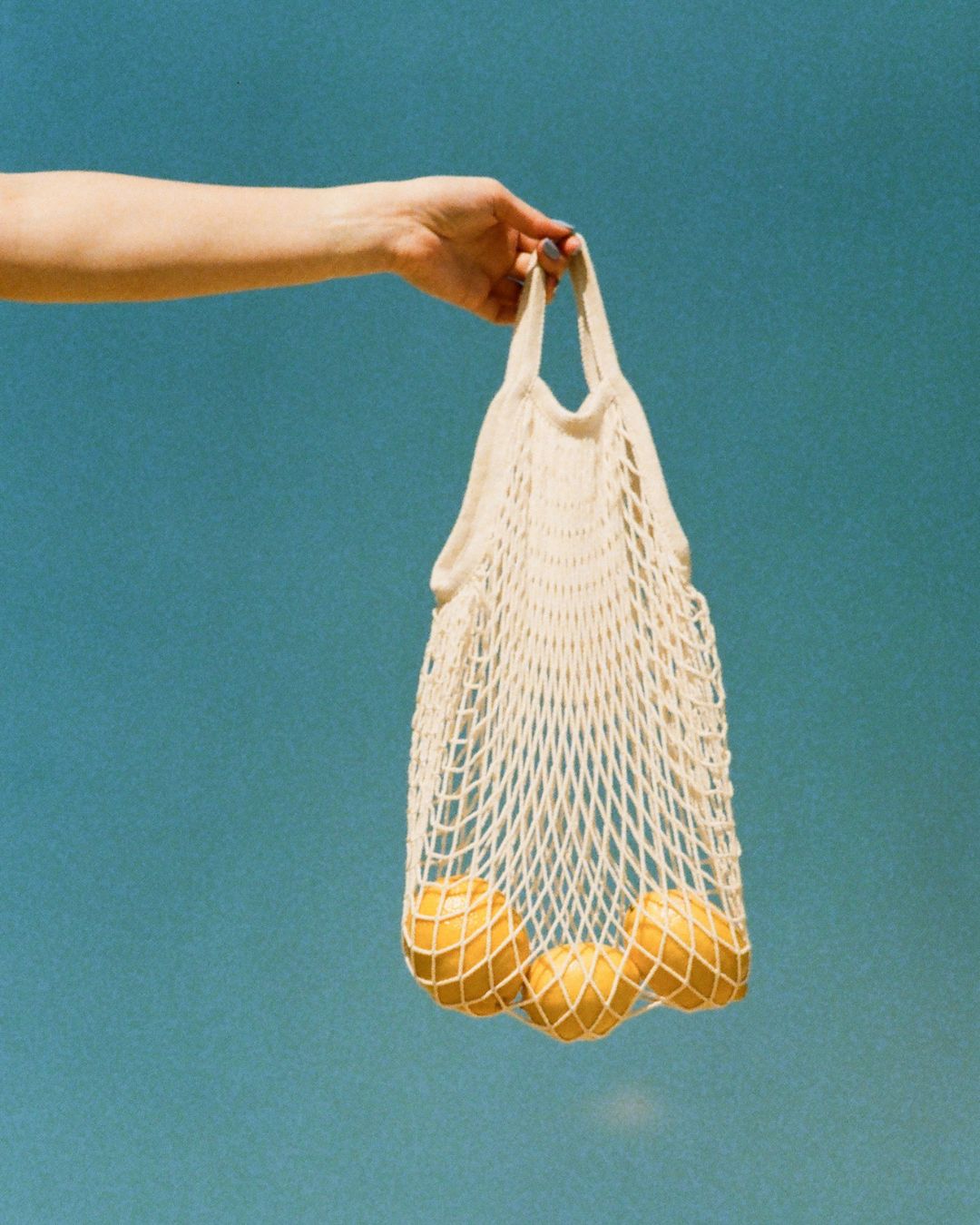 Hand holding an ECOBAGS natural undyed cotton string net bag with lemons against a blue sky background