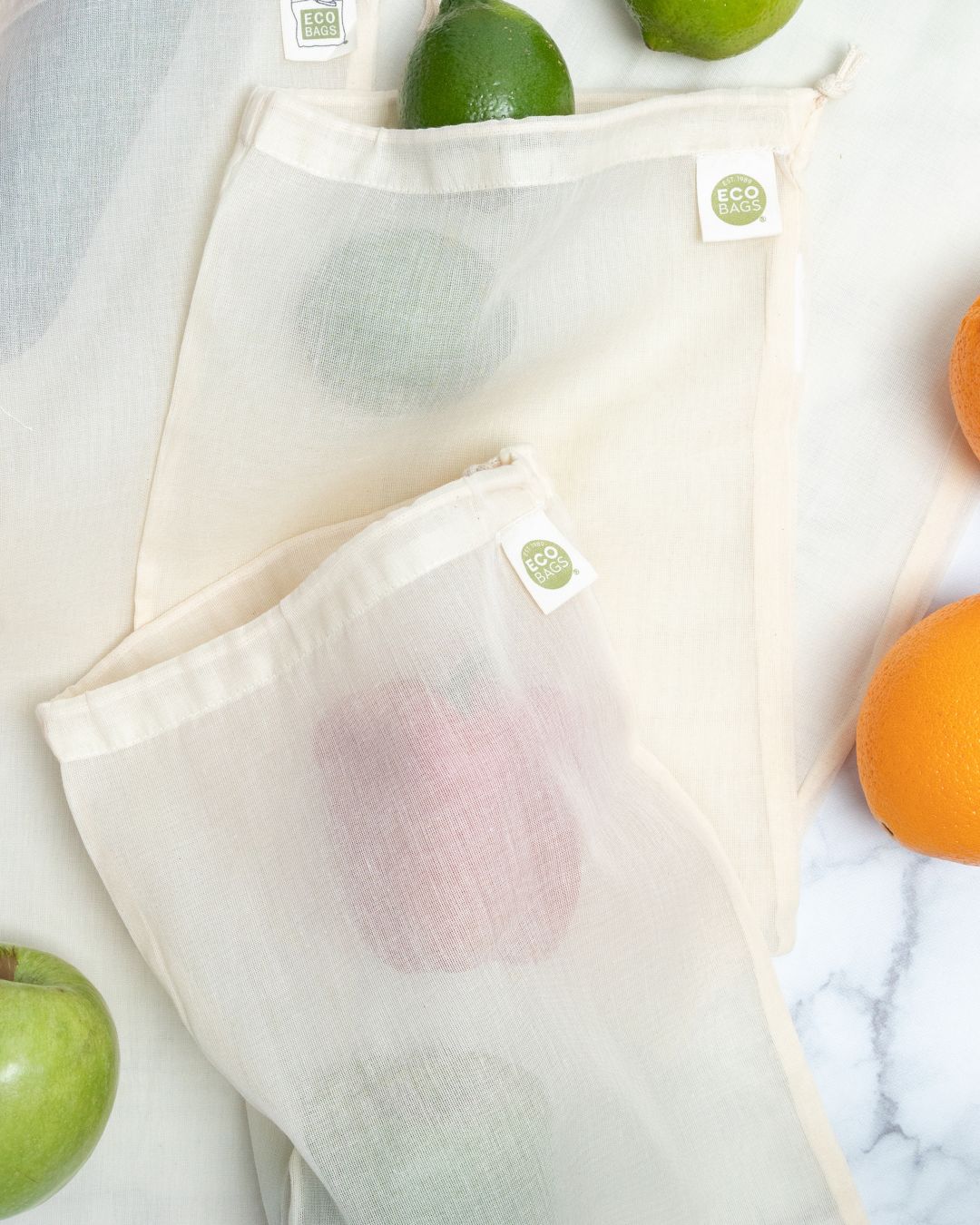 Reusable gauze produce bags with a visible green ecobags logo containing fruits on a marble surface