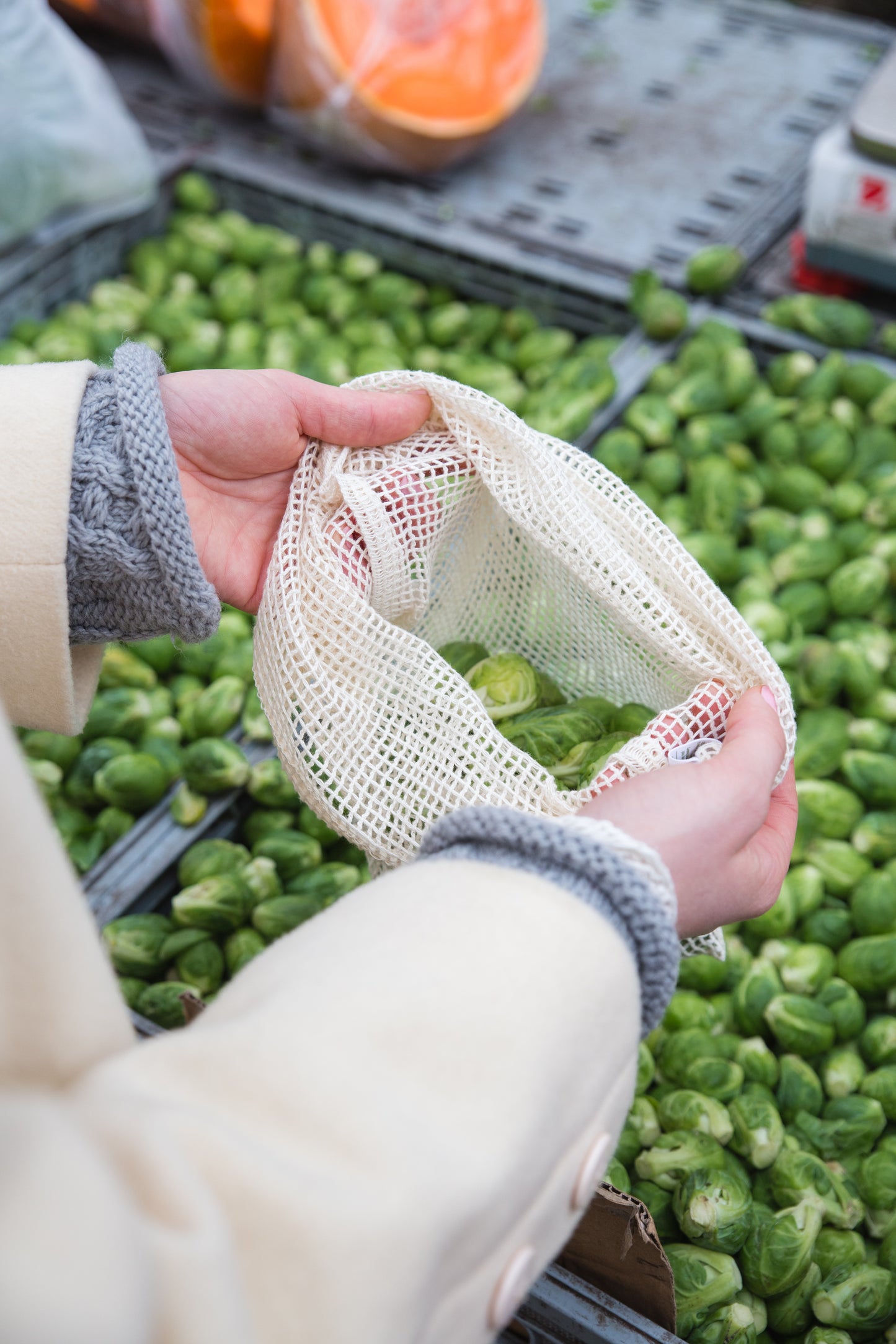 A person holds a natural colored, cotton mesh bag containing brussel sprouts. Behind them are flats of more veggies.