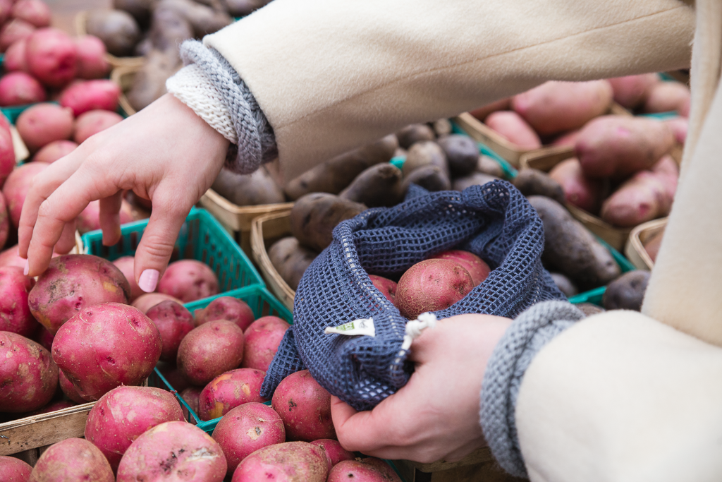 A hand reaches for a small potato while holding a mesh bag  full of other produce