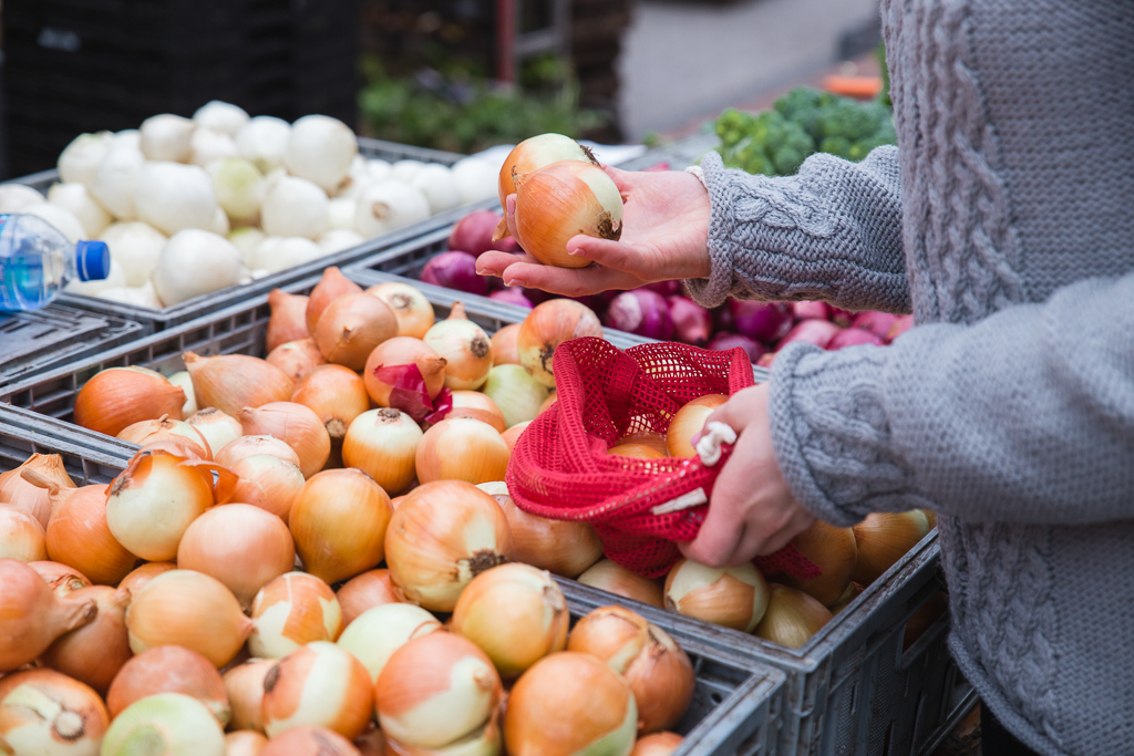A person holds onions in one hand and a bright red mesh sack full of them in the other while at a farmer's market
